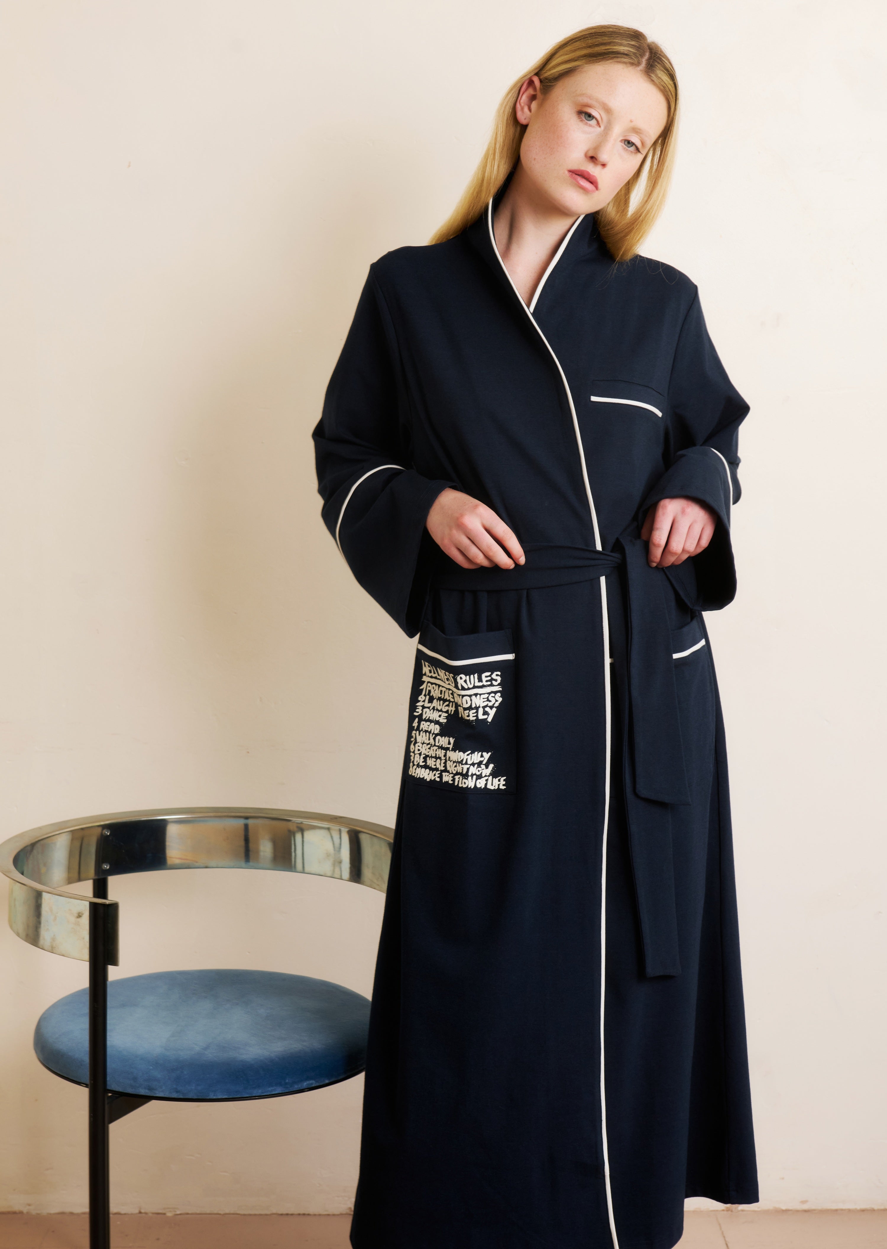Woman wearing a long black coat with white text and piping in a minimalistic room.